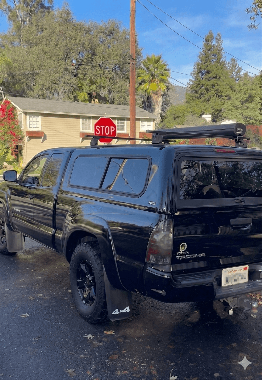 Black Toyota Tacoma with matching camper shell, roof rack, and 4x4 mud flaps on a residential street